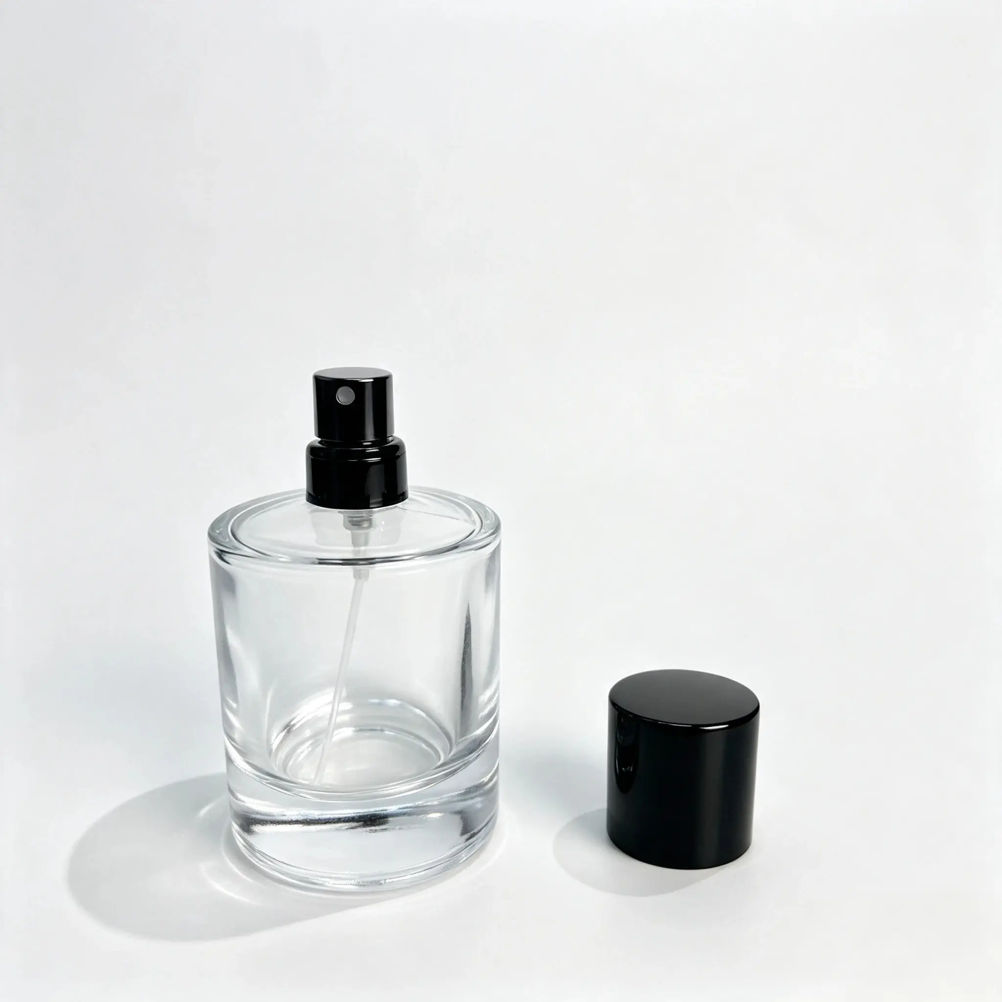 100ml clear cylindrical glass perfume bottle shown bare with the crimp sprayer installed and outer cap standing separately beside it
