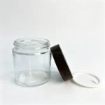 100g clear glass cream jar displayed with its separate wood-grain screw cap, showing the container and closure components for facial cream packaging.