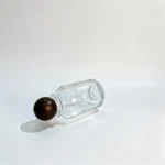 Side-lying view of the 100ml round perfume bottle with its wooden ball cap displayed separately, showing the relationship between the bulbous bottle body and spherical cap.