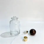 Uncapped 100ml round perfume bottle with the wooden ball cap, fragrance sprayer and dip tube arranged separately beside the bottle to show its packaging components.