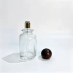Uncapped 100ml round perfume bottle with its wooden ball cap placed beside the bottle, showing the rounded bottle profile and removable cap as separate components.