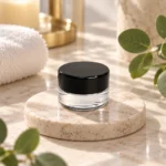 Small clear glass cream sample jar with a black lid displayed in a skincare packaging setting.