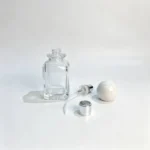 Separate perfume sprayer, dip tube, and white ball cap components arranged beside the uncapped 50ml square fragrance bottle.