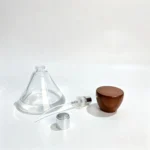 Uncapped 50ml wide round perfume bottle displayed beside its fragrance sprayer, dip tube, and conical wooden cap for an overview of the separate packaging components.